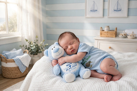 A sleeping newborn baby in a light blue onesie rests on a white textured blanket, hugging a blue plush toy with personalized text. The baby is posed in a bright, sunlit nursery with light blue and white striped walls, framed sailboat prints, and a white dresser in the background.