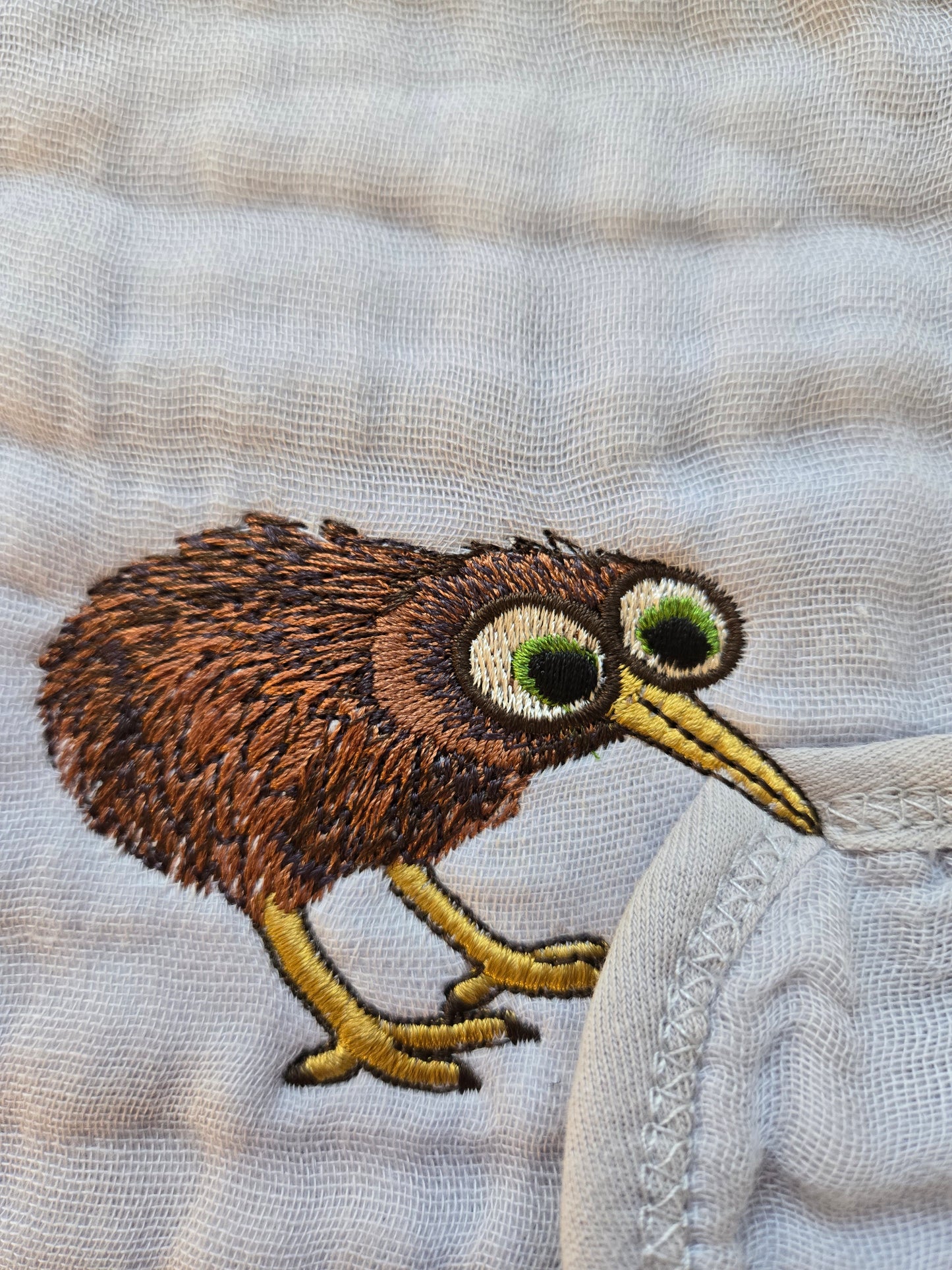 Close up of high density embroidery featuring a funny Kiwi bird stealing a pavlova cake on a sage green kitchen tea towel.