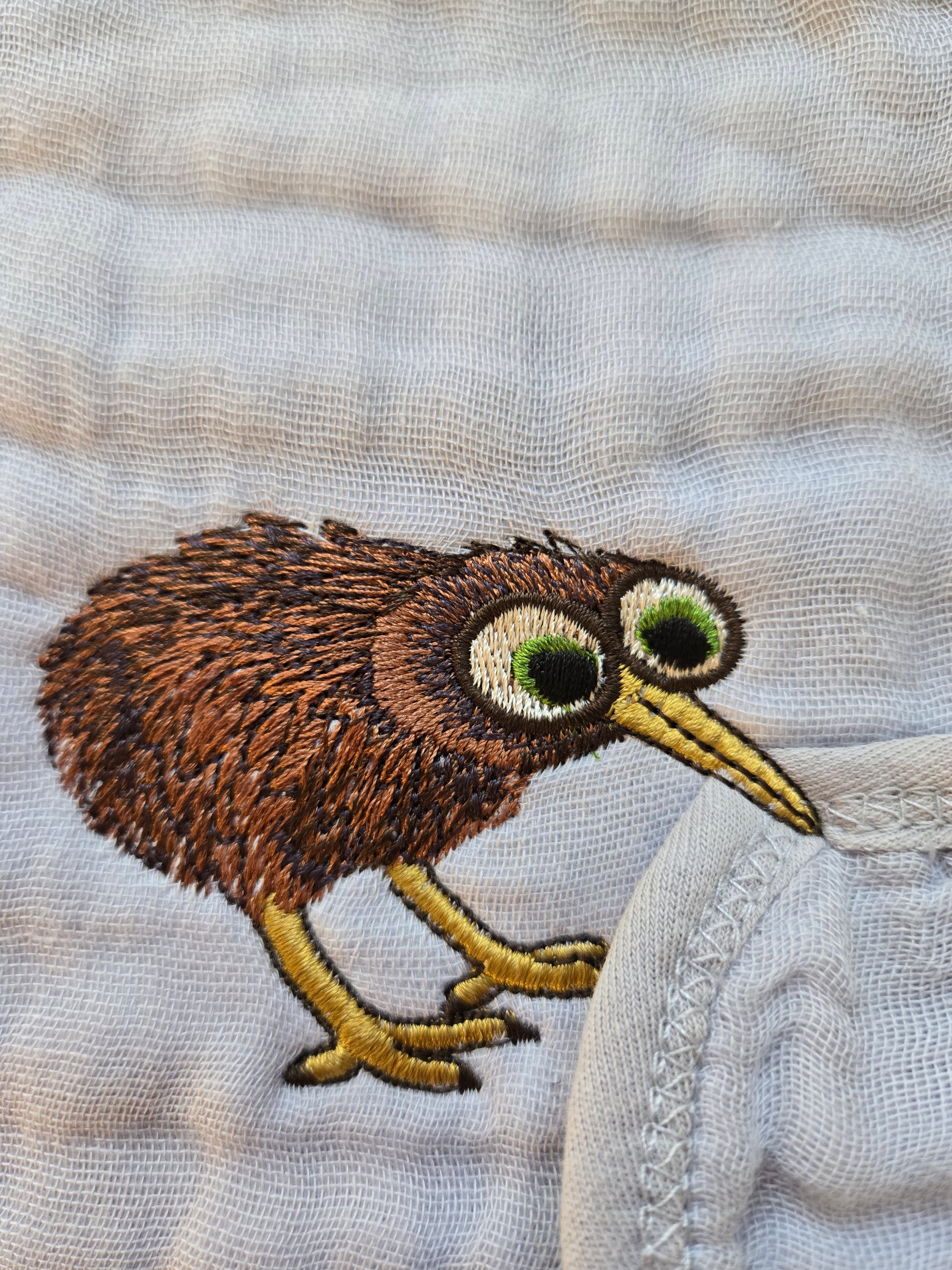 Close up of high density embroidery featuring a funny Kiwi bird stealing a pavlova cake on a sage green kitchen tea towel.