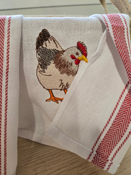 A white linen tea towel with red striped borders and an embroidered brown, black, red, and yellow chicken on the front, draped over a wooden surface.