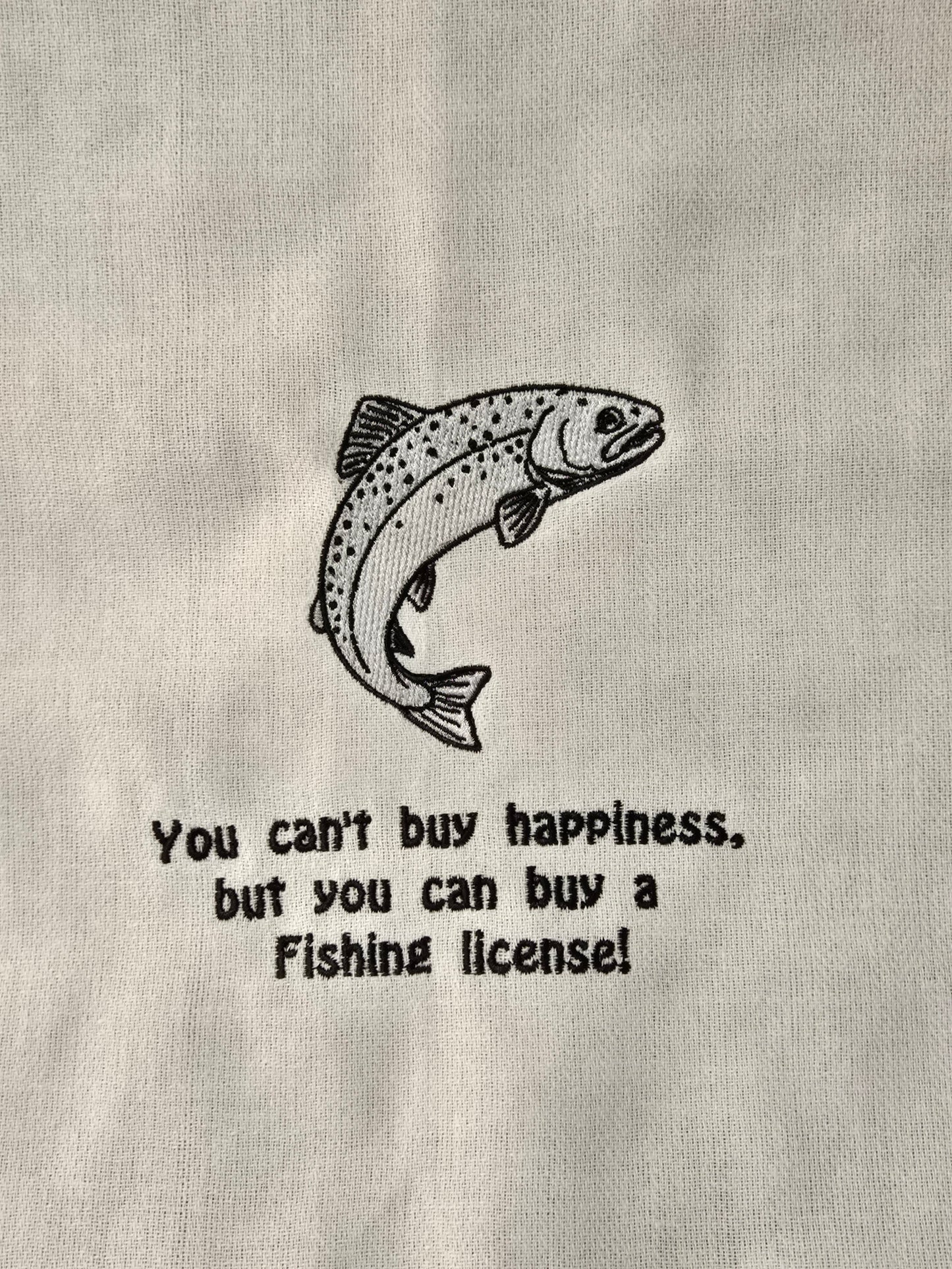 A close-up of a light gray fabric surface with a black line drawing of a jumping fish and the text, 'You can't buy happiness, but you can buy a Fishing license!' printed below it.