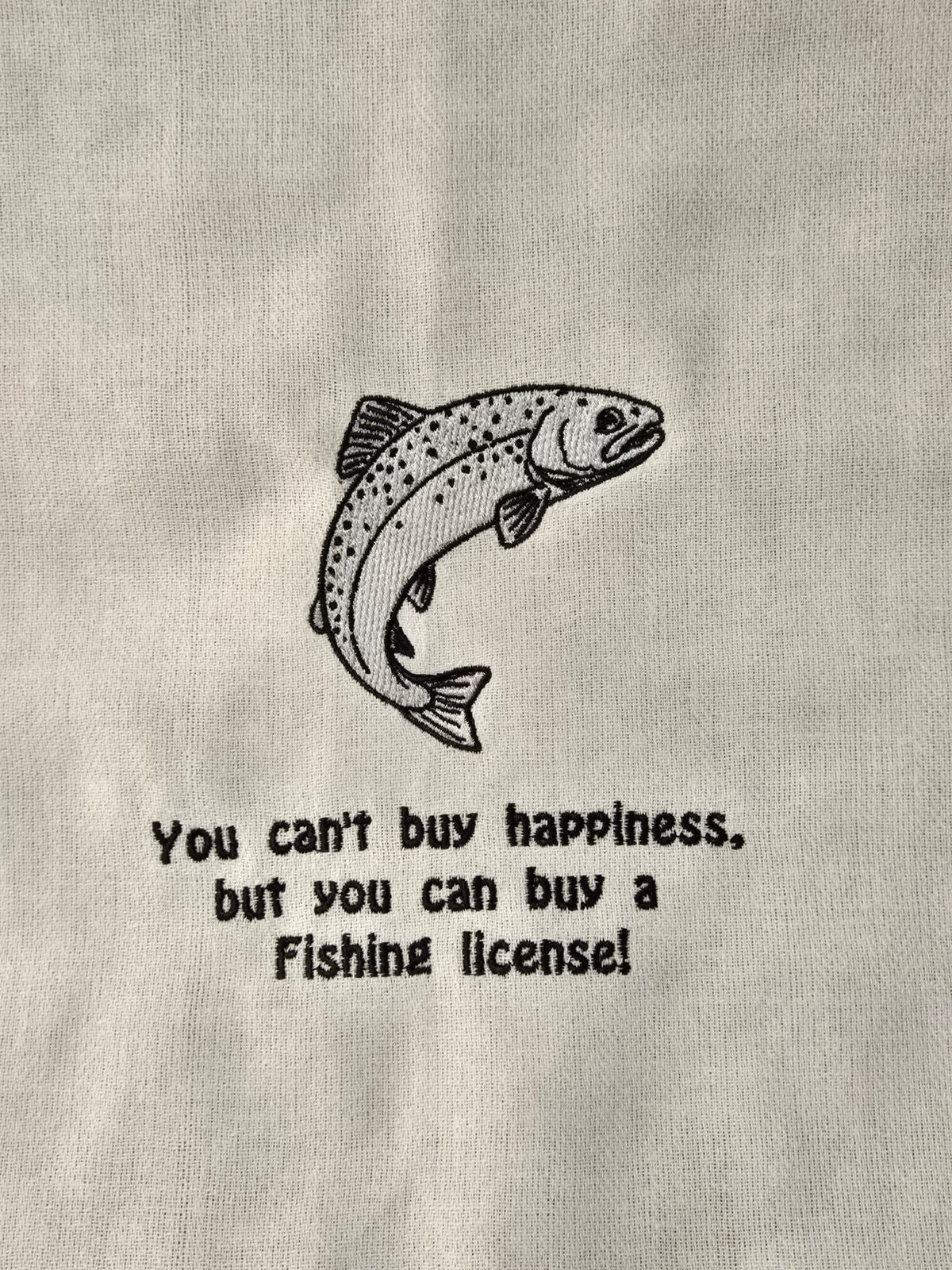 A close-up of a light gray fabric surface with a black line drawing of a jumping fish and the text, 'You can't buy happiness, but you can buy a Fishing license!' printed below it.