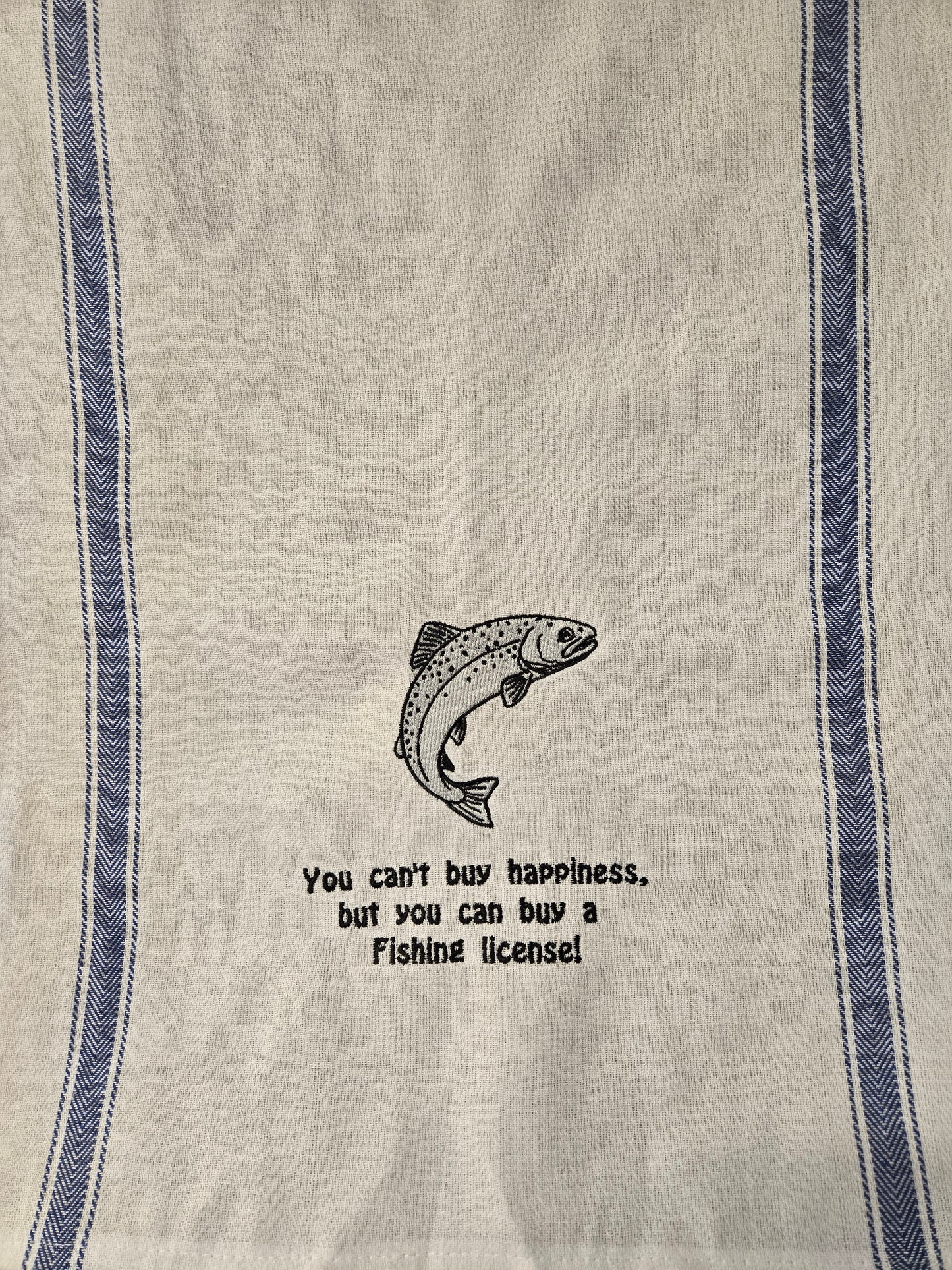 A close-up of a woven kitchen towel or tea towel in a warm off-white linen fabric, featuring two vertical blue stripes and a central black graphic of a jumping fish with the text 'You can't buy happiness, but you can buy a Fishing license!'.