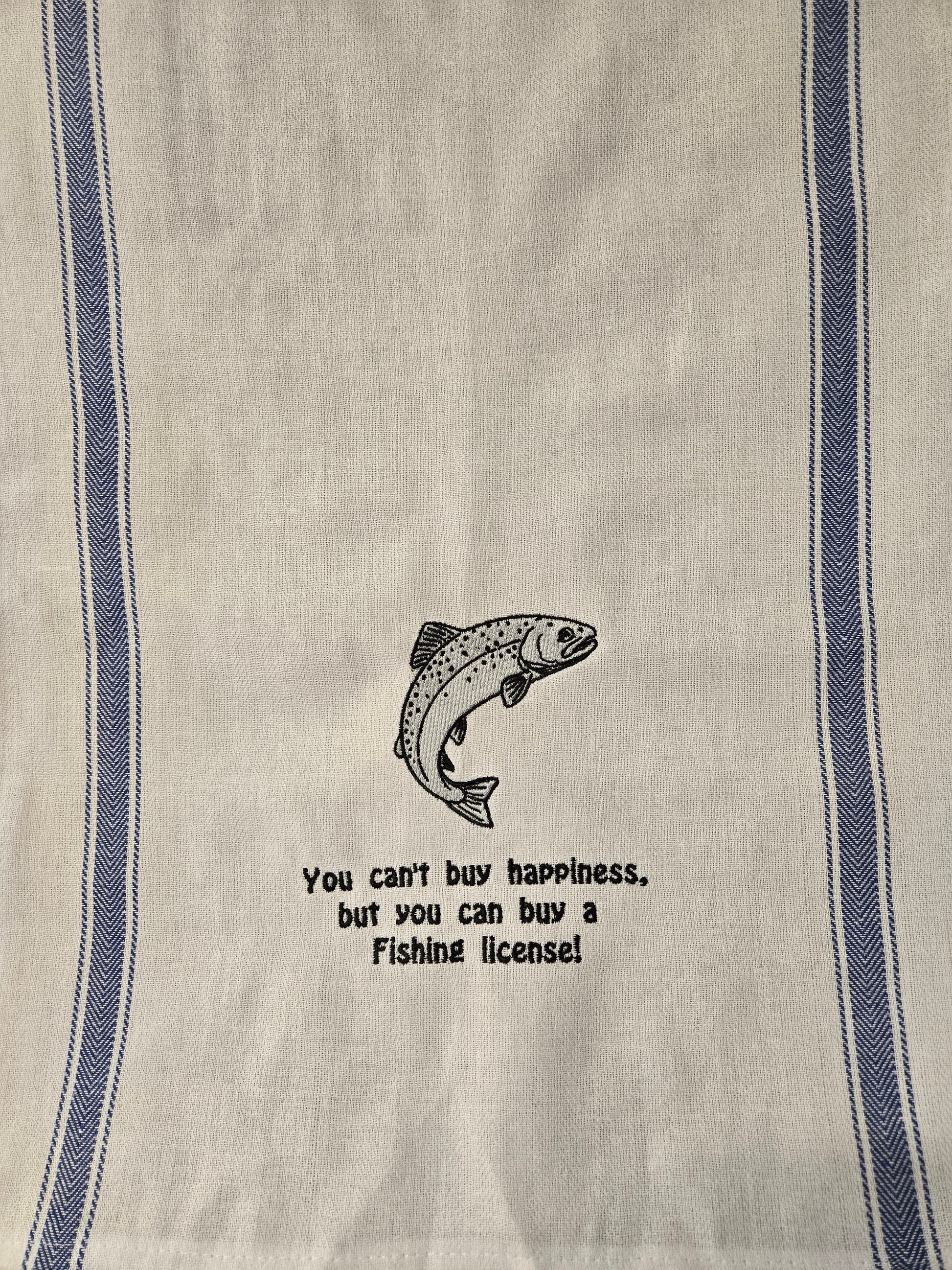 A close-up of a woven kitchen towel or tea towel in a warm off-white linen fabric, featuring two vertical blue stripes and a central black graphic of a jumping fish with the text 'You can't buy happiness, but you can buy a Fishing license!'.