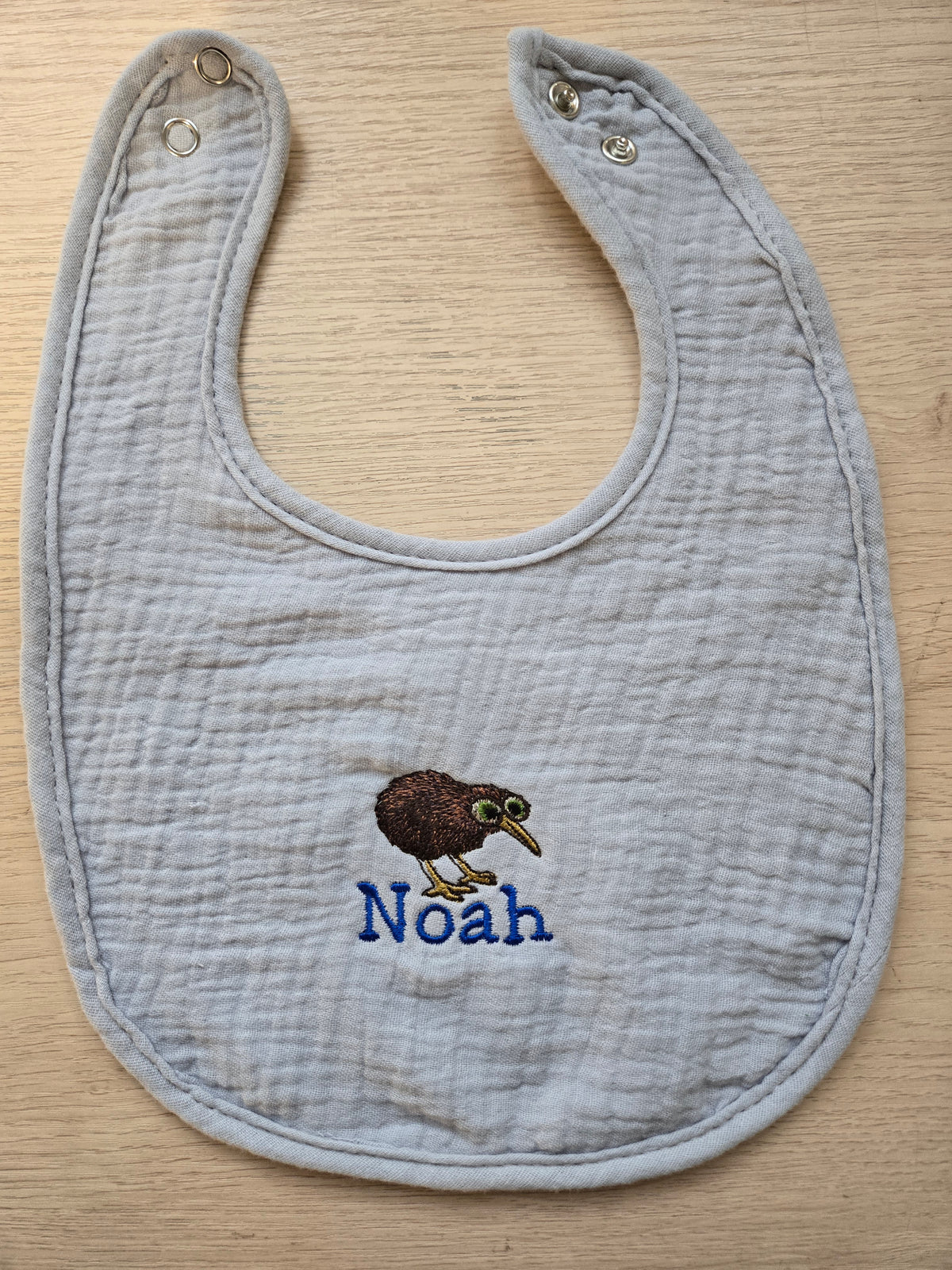 Gray baby bib with embroidered kiwi bird and 'Noah' text on a wooden surface