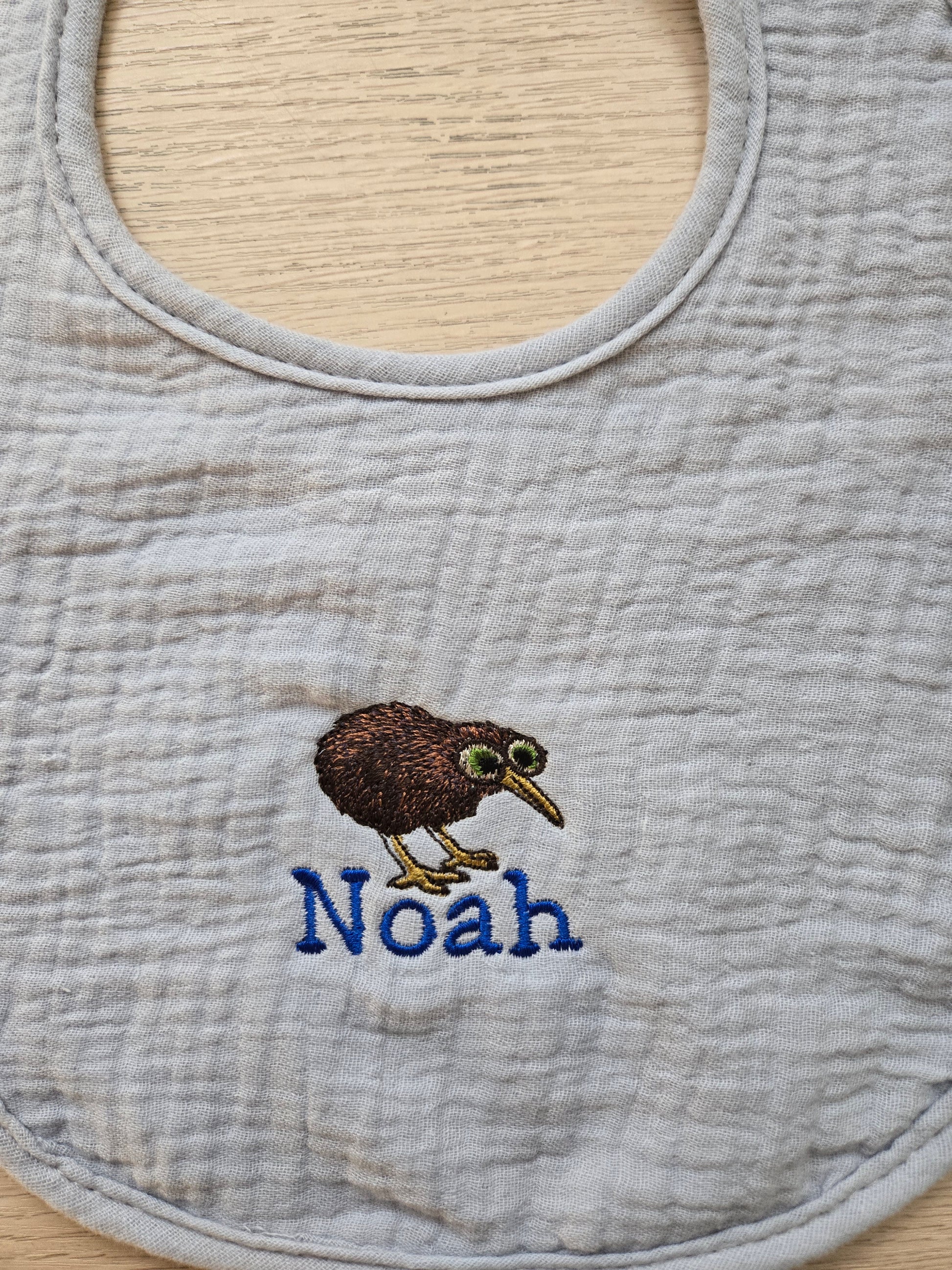Gray bib with embroidered kiwi bird and 'Noah' text on a wooden surface