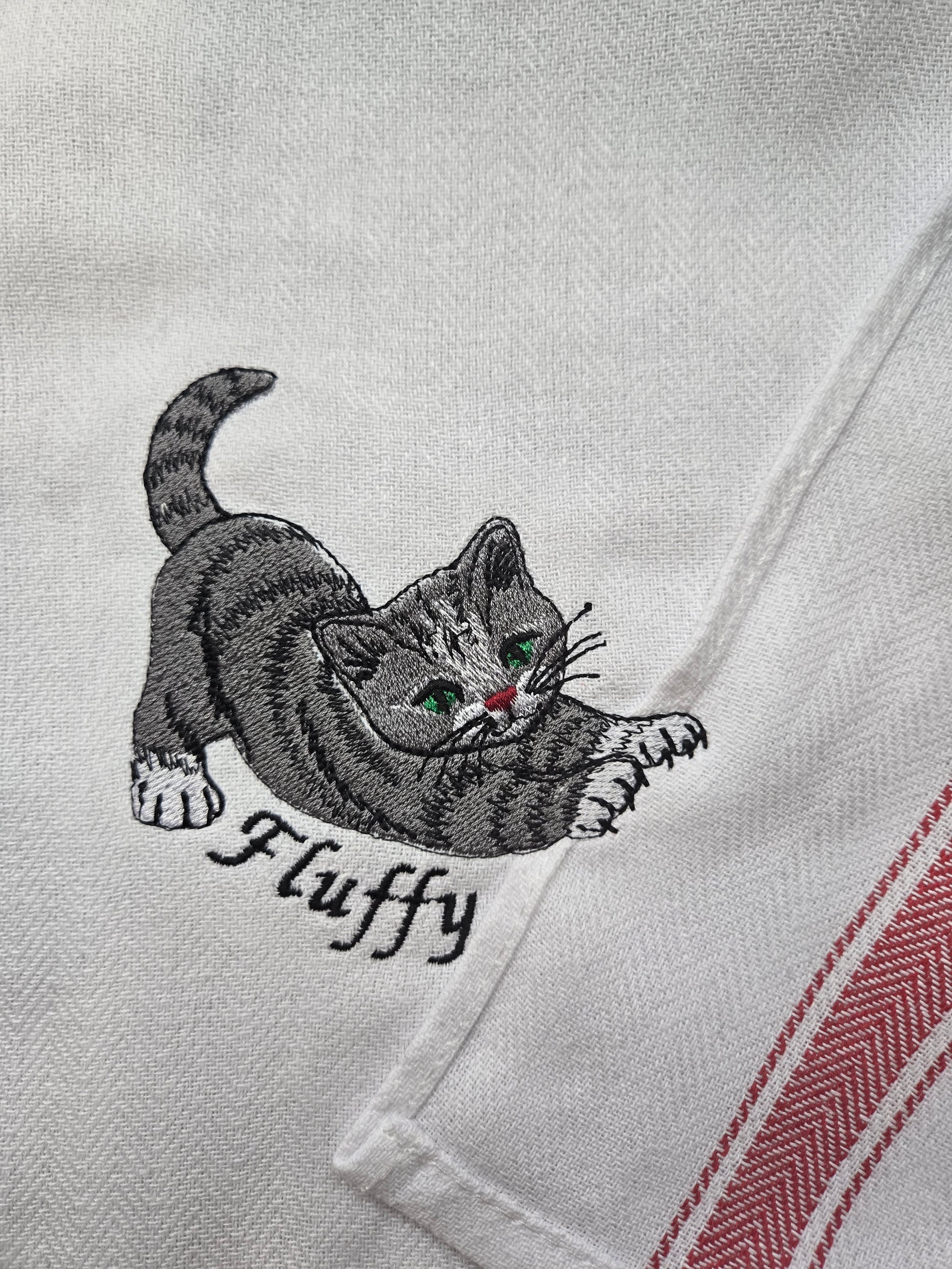 Close-up of a white embroidered tea towel featuring a gray tabby kitten with green eyes and the name 'Fluffy' embroidered underneath, with red and white herringbone stripes on the right edge.