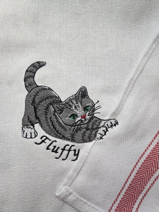 Close-up of a white embroidered tea towel featuring a gray tabby kitten with green eyes and the name 'Fluffy' embroidered underneath, with red and white herringbone stripes on the right edge.