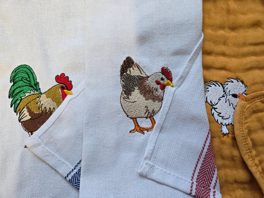 A close-up of folded white kitchen towels featuring embroidered farm animals, including a rooster, hen, and chick, with a mustard yellow quilted blanket partially visible on the right.
